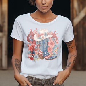 May include: White t-shirt featuring a watercolor design of a cowboy hat, boots, and saddle adorned with flowers. The boots display a patriotic American flag design with stars and stripes. The design incorporates pink, blue, and red floral elements.