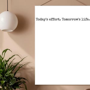 以下が含まれることがあります： 黒い文字で「Today's effort. Tomorrow's life.」と書かれた長方形の白いウォールアートプリント。プリントは縦向きで、上端に取り付けられた細い紐で薄茶色の壁面に吊り下げられています。