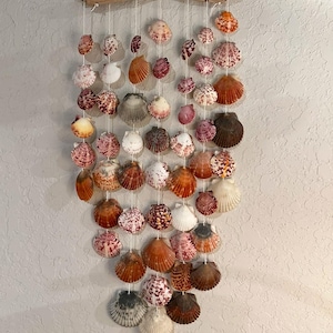 May include: A seashell wind chime made of various shades of brown, white, and pink seashells hanging from a driftwood branch.