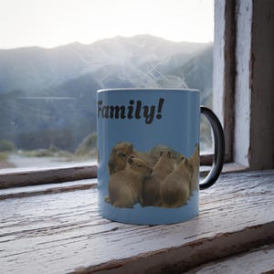 Puede incluir: Taza de cerámica azul con la palabra "Family!" en escritura negra. La taza presenta un grupo de carpinchos. La taza tiene un asa negra y está colocada frente a una ventana con vista a la montaña.