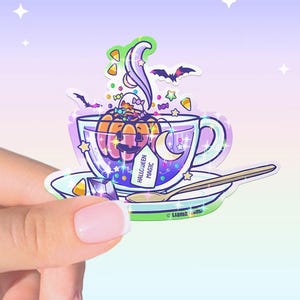 May include: A holographic sticker featuring a clear teacup filled with purple liquid, a smiling orange pumpkin, candy corn, stars, and bats. A tea tag reads HALLOWEEN MAGIC. The cup sits on a saucer with a spoon, all accented by sparkles.