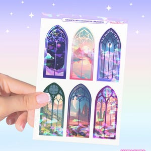 May include: A sticker sheet titled Cathedral Windows Ethereal Landscapes featuring six arched window designs. Each window displays a unique scene, including space, mountains, and desert landscapes with vibrant, iridescent, and colorful details.