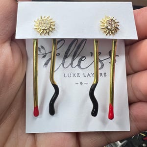 Puede incluir: Un par de pendientes de oro con un diseño negro y rojo. Los pendientes presentan un pequeño sol con brillantes transparentes. Los pendientes se muestran en una tarjeta blanca con el texto "Elle's Luxe Layers" impreso en ella.