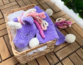 Puede incluir: Una manta de unicornio de crochet morado con un pompón blanco y detalles de hilo rosa y blanco. La manta está en una cesta de mimbre.
