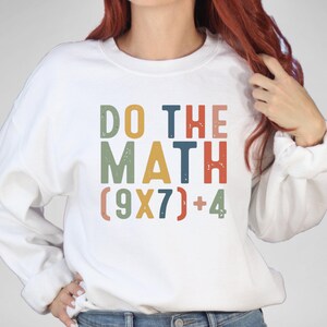 May include: White sweatshirt with the text "DO THE MATH" in colorful, distressed lettering. Below the text is the equation (9X7)+4. The sweatshirt has long sleeves and a crew neck.