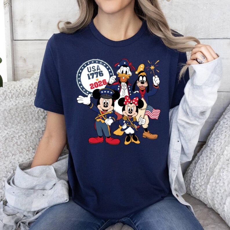 May include: Navy blue t-shirt featuring a patriotic design with Mickey Mouse, Minnie Mouse, Donald Duck, and Goofy in red, white, and blue outfits. The shirt includes the text "USA 1776 2026" in a circular design.