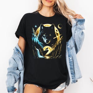 May include: Black t-shirt featuring a wolf design. The graphic depicts a wolf's face against a full moon, with a smaller wolf howling at a moonlit landscape. The design incorporates elements of nature, including trees and a waterfall.