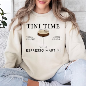 May include: Cream-colored sweatshirt with the text "TINI TIME" above an illustration of an espresso martini. The ingredients, vodka, espresso, and coffee liqueur, are listed. The sweatshirt is a casual, comfortable garment.