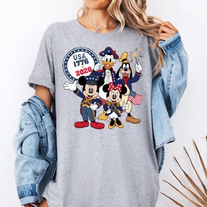 May include: A gray t-shirt featuring a patriotic design with Mickey Mouse, Minnie Mouse, Donald Duck, and Goofy in red, white, and blue outfits. The shirt includes the text "USA 1776 2026" in a circular emblem.