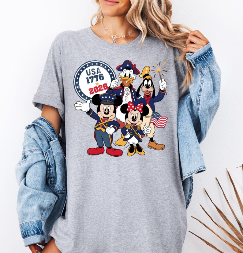 May include: A gray t-shirt featuring a patriotic design with Mickey Mouse, Minnie Mouse, Donald Duck, and Goofy in red, white, and blue outfits. The shirt includes the text "USA 1776 2026" in a circular emblem.