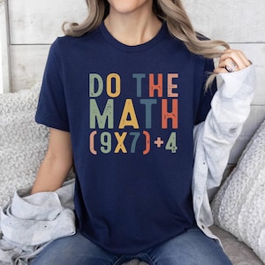 May include: Navy blue t-shirt with the text "DO THE MATH (9X7)+4" in colorful, distressed lettering. The shirt has a classic crew neck and short sleeves. The design features orange, green, blue, and red text.