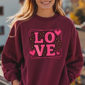 May include: Burgundy sweatshirt with a pink "LOVE" graphic. The letters have leopard print and polka dot patterns. Pink hearts frame the text, creating a Valentine's Day theme.