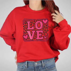 May include: Red sweatshirt with the word "LOVE" in pink letters. The letter "O" has a zebra stripe pattern, and the letters "L", "V", and "E" have a pink heart design. The background has a leopard print.