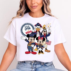 May include: White t-shirt featuring a patriotic design with Mickey Mouse, Minnie Mouse, Donald Duck, and Goofy in red, white, and blue outfits. The design includes the text "USA 1776 2026" in a circular emblem, celebrating American Independence.