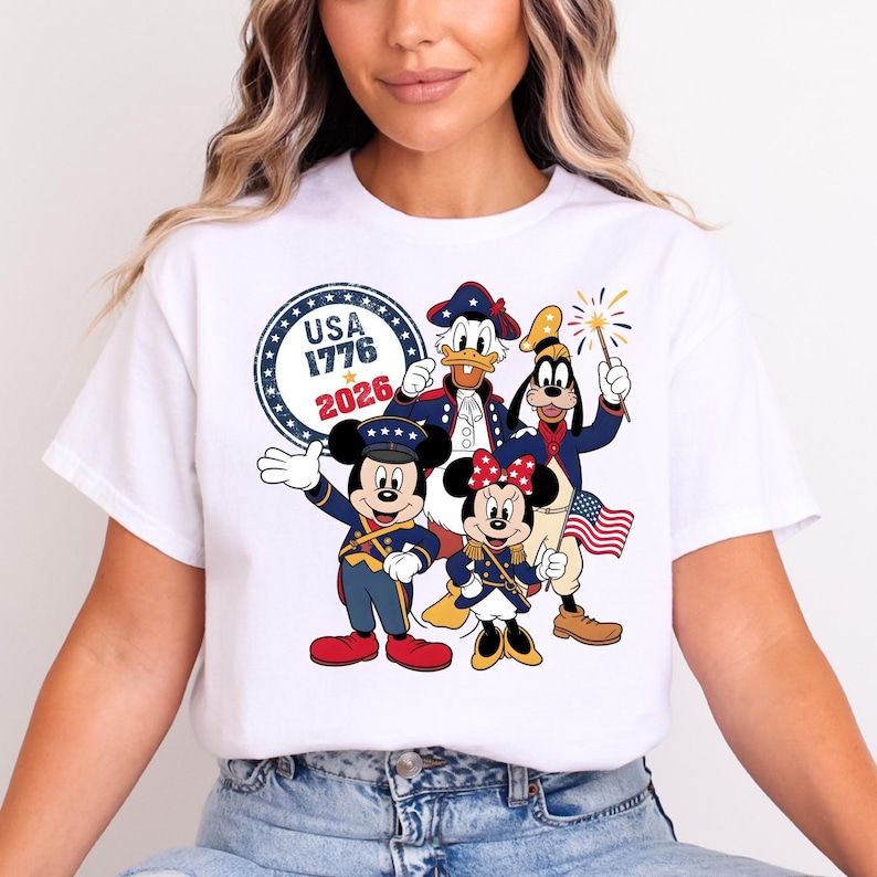 May include: White t-shirt featuring a patriotic design with Mickey Mouse, Minnie Mouse, Donald Duck, and Goofy in red, white, and blue outfits. The design includes the text "USA 1776 2026" in a circular emblem, celebrating American Independence.