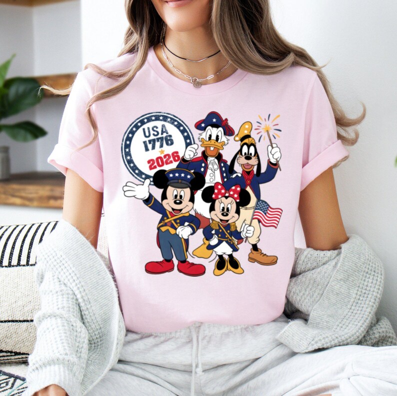 May include: A light pink t-shirt featuring a patriotic design with cartoon characters. The graphic includes Mickey Mouse, Minnie Mouse, Donald Duck, and Goofy in colonial-style outfits, along with the text "USA 1776 2026" in a circular emblem.