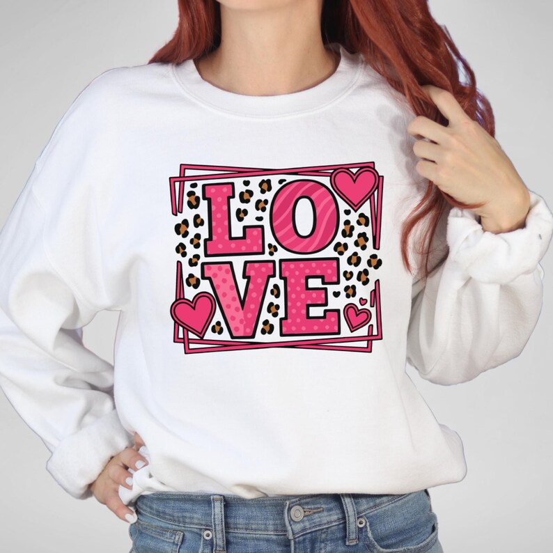 May include: White sweatshirt with a pink and leopard print "LOVE" graphic. The word "LOVE" is in a pink outlined box with pink hearts. The letter "O" has a zebra stripe pattern. The sweatshirt is a casual style.