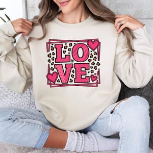 May include: Cream-colored sweatshirt with a pink "LOVE" graphic. The letters are outlined in pink and filled with leopard and zebra print patterns. Pink hearts are placed around the letters.