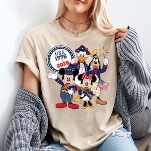 May include: Beige t-shirt featuring a patriotic design with Mickey Mouse, Minnie Mouse, Donald Duck, and Goofy in red, white, and blue outfits. The shirt includes the text "USA 1776 2026" in a circular emblem, celebrating American Independence.