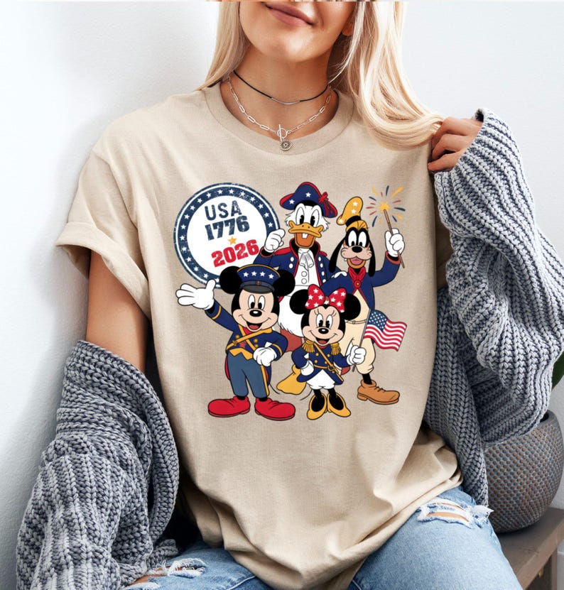 May include: Beige t-shirt featuring a patriotic design with Mickey Mouse, Minnie Mouse, Donald Duck, and Goofy in red, white, and blue outfits. The shirt includes the text "USA 1776 2026" in a circular emblem, celebrating American Independence.