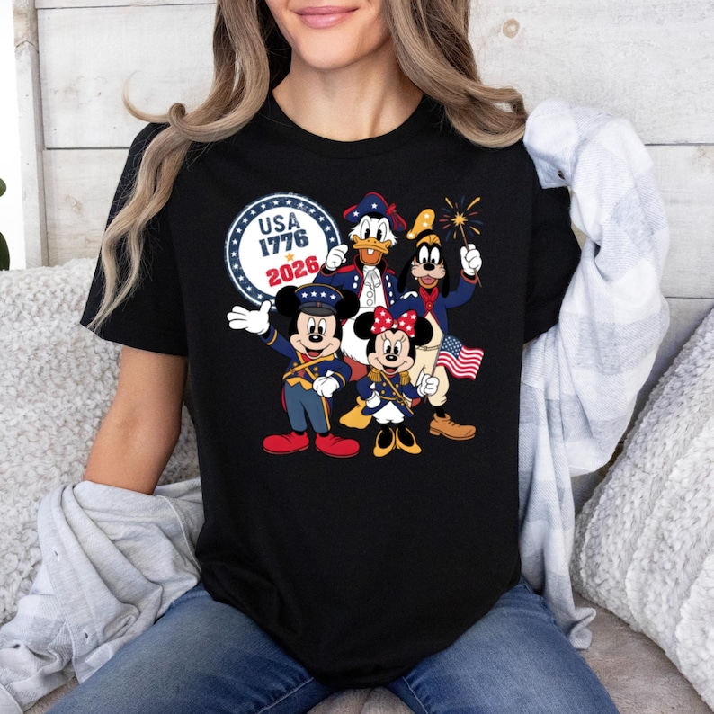 May include: Black t-shirt featuring a patriotic design with Mickey Mouse, Minnie Mouse, Donald Duck, and Goofy in red, white, and blue outfits. The shirt includes the text "USA 1776 2026" in a circular emblem.