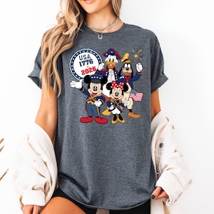 May include: Dark gray t-shirt featuring a graphic of Mickey Mouse, Minnie Mouse, Donald Duck, and Goofy in patriotic attire. The design includes the text "USA 1776 2026" in a circular emblem, with fireworks and an American flag.
