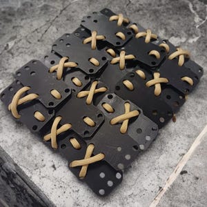 May include: A close-up of a set of black metal buckles with tan straps woven through the holes. The buckles are arranged in a staggered pattern.