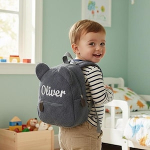 May include: A gray teddy bear-shaped backpack with the name "Oliver" embroidered in white. The backpack has two small ear-shaped details and brown zipper pulls. The child is wearing a striped shirt and khaki pants.