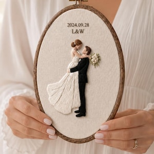 May include: An oval embroidery hoop containing a fabric panel with a stitched illustration of a couple embracing. The bride wears a white gown and the groom wears a black suit. Above them, the text 2024.09.28 and L&W is embroidered in brown.