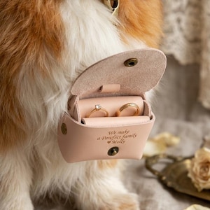 May include: A light pink leather ring box with a snap closure and a gold-tone metal ring holder inside. The box is engraved with the text "We make a Pawfect family Molly" and contains two gold rings, one with a small clear stone.