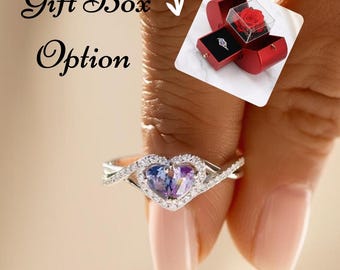 May include: A silver-toned ring featuring a heart-shaped center stone with blue and purple hues, surrounded by small clear stones on a split band. An inset shows a red gift box containing a ring and a preserved red rose, labeled Gift Box Option.