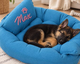 May include: A bright blue pet bed with a raised back cushion featuring the embroidered pink name Max and a pink paw print icon. A black and tan German Shepherd puppy is curled up asleep in the center of the soft, textured fabric bed.