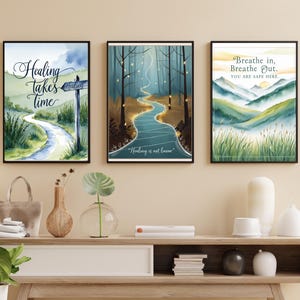 Op de afbeelding: Drie ingelijste aquarellen met natuurscènes en inspirerende citaten. De eerste schildering toont een kronkelend pad door een groene weide met de quote "Genezing kost tijd". De tweede schildering toont een blauwe rivier die door een bos slingert met de quote "Genezing is niet lineair". De derde schildering toont een bergketen met de quote "Adem in, adem uit. Je bent hier veilig."