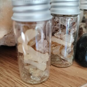 May include: Two small, clear glass vials with silver-colored screw-top lids. Each vial is filled with a collection of translucent, pale yellow and off-white resinous chunks or crystal-like fragments, appearing irregular in shape and size.