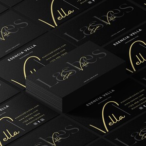 Könnte beinhalten: Ein Stapel schwarzer Visitenkarten mit goldener Schrift und einem goldenen Logo. Die Karten zeigen den Namen "Esencia Vella Lashes" zusammen mit Kontaktinformationen. Die Karten sind auf einer dunklen Oberfläche angeordnet.