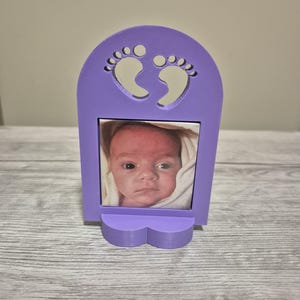 May include: A lilac picture frame holding a baby's photograph. The frame features a cutout of two footprints at the top and a heart-shaped base. The photo shows a baby wrapped in a white blanket.