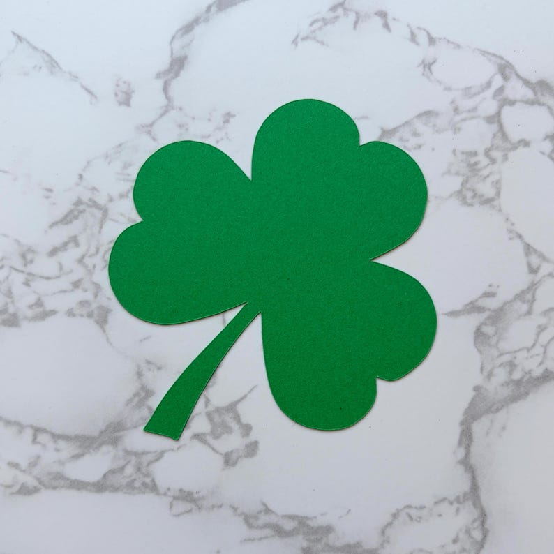 May include: A bright green shamrock with three leaves and a stem. The clover is cut out of a solid green material and is set against a white and gray marble background. A symbol of St. Patrick's Day.