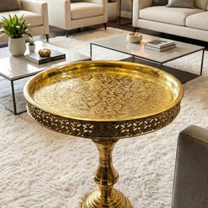 May include: A gold-coloured, ornate coffee table with a round, engraved tray top and a decorative pedestal base. The table is on a white shag rug in a living room setting.