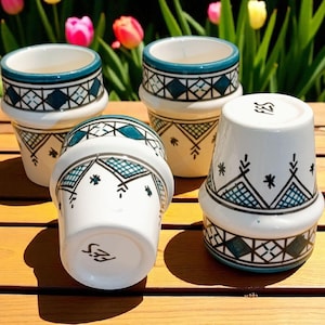 May include: Four white ceramic cups with teal and black geometric patterns. The cups have a flared shape with a teal rim and a decorative band around the base. The cups are arranged on a wooden surface.