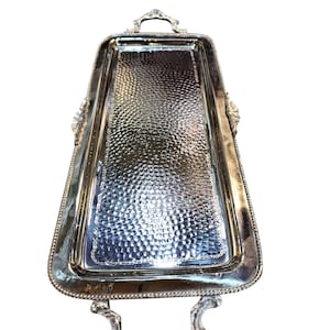 May include: A rectangular silver serving tray with ornate handles and a textured surface. The tray has a decorative border and is designed for serving food or drinks. The handles are curved and add to the tray's elegant design.