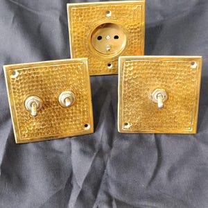 Könnte beinhalten: Drei goldfarbene elektrische Armaturen mit gehämmertem Muster. Das Set umfasst eine Steckdose und zwei Lichtschalter. Die Armaturen sind quadratisch mit abgerundeten Ecken und haben eine Vintage-Ästhetik.