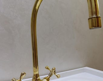 May include: A gold-colored faucet with a curved spout and cross-shaped handles. The faucet is made of a shiny metal and is set against a white surface and a neutral-toned wall. The design is classic and elegant.