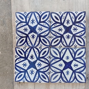 Puede incluir: Cuatro baldosas de cerámica cuadradas con fondo blanco y diseño floral azul. Cada baldosa presenta una forma de estrella central rodeada de hojas estilizadas y pequeños acentos de estrella. Las baldosas están dispuestas sobre una superficie de madera.
