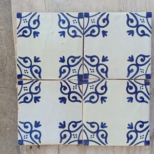 Może przedstawiać: Cztery kwadratowe płytki ceramiczne z białym tłem i powtarzającym się niebieskim wzorem kwiatowym. Płytki są ułożone razem, prezentując wzór. Wzór zawiera wirujące linie i stylizowane elementy kwiatowe, tworząc dekoracyjną powierzchnię.