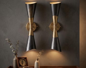 May include: Two hourglass-shaped wall sconces with a black and brass finish. The sconces are mounted on a dark gray wall, casting a warm glow. The sconces are part of a home decor theme.