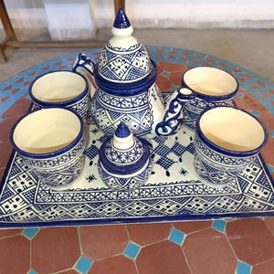 May include: A white and blue ceramic tea set with a teapot, sugar bowl, and four cups. The set features a geometric pattern in blue. The tea set is on a rectangular tray with a matching design.