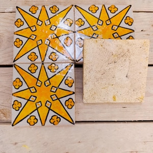 Op de afbeelding: Vier decoratieve keramische tegels met een sterrenpatroon in geel en zwart op een witte achtergrond. De tegels zijn gerangschikt met een enkele, ongeverfde tegel. De tegels liggen op een licht houten oppervlak.