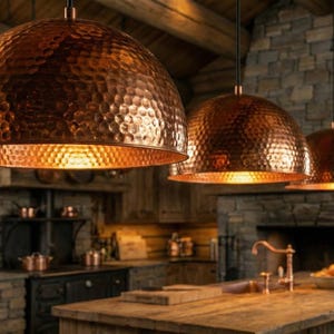 May include: Three copper pendant lights with a hammered texture hang above a rustic kitchen. The lights have a warm, orange glow, complementing the wooden cabinets and stone fireplace. The kitchen features a wooden island and a stove.