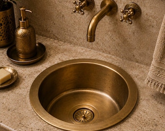 May include: A round, antique brass bathroom sink basin with a matching wall-mounted faucet featuring two cross-handle knobs. A decorative brass soap dispenser sits on a small tray next to the sink, which is set into a light-colored stone countertop.