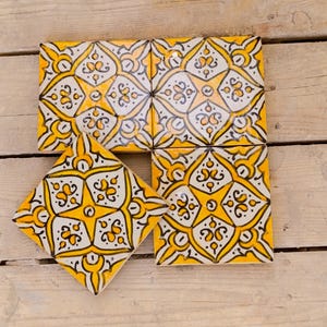 May include: A collection of yellow, white, and black decorative tiles. The tiles feature a repeating star and floral pattern. The tiles are square and arranged on a wooden surface. The tiles are likely for home decor or craft projects.